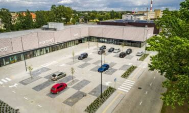 Mažos automobilių stovėjimo aikštelės prie modernios prekyboscentro eurokos, su žaliaisais medžiais ir pėsčiųjų perėjomis, aplinkui gamta ir gyvenamieji namai, svarbi tema apie miesto gyvenamąją aplinką ir parduotuvių infrastruktūrą.