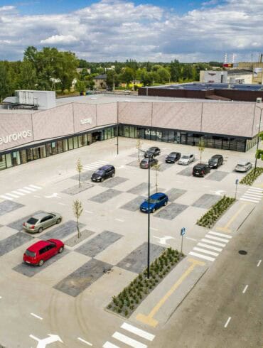 Mažos automobilių stovėjimo aikštelės prie modernios prekyboscentro eurokos, su žaliaisais medžiais ir pėsčiųjų perėjomis, aplinkui gamta ir gyvenamieji namai, svarbi tema apie miesto gyvenamąją aplinką ir parduotuvių infrastruktūrą.